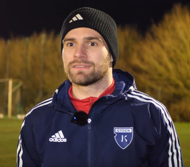 Ein Mann in einer blauen Jacke und Mütze auf einem Fußballfeld während eines Trainings, mit Bäumen und einem Tor im Hintergrund.