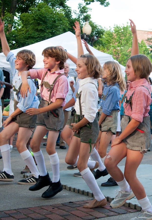 Eine Gruppe von Kindern in traditioneller bayrischer Tracht, die auf einer Straße tanzen, umgeben von einer Menge, Zelten, Laternenmasten, Bäumen, Gebäuden und einem klaren blauen Himmel, wobei einige Kinder Shorts und Schuhe tragen.