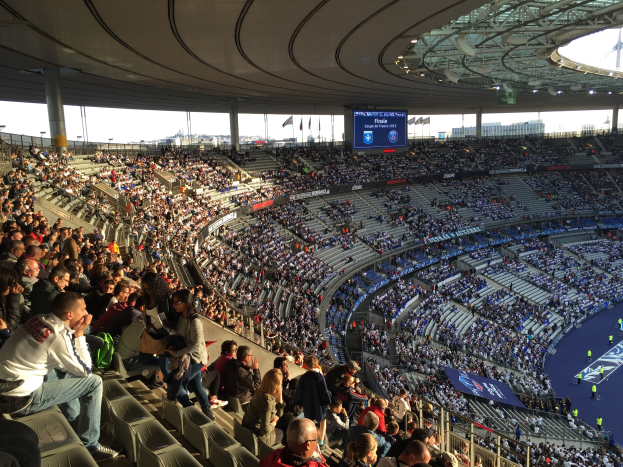 Große Zuschauermenge in einem Stadion bei einem Fußballspiel, mit einer Bühne rechts, Fahnen, Masten, einem Bildschirm und der Allianz Arena in München, Deutschland im Hintergrund.