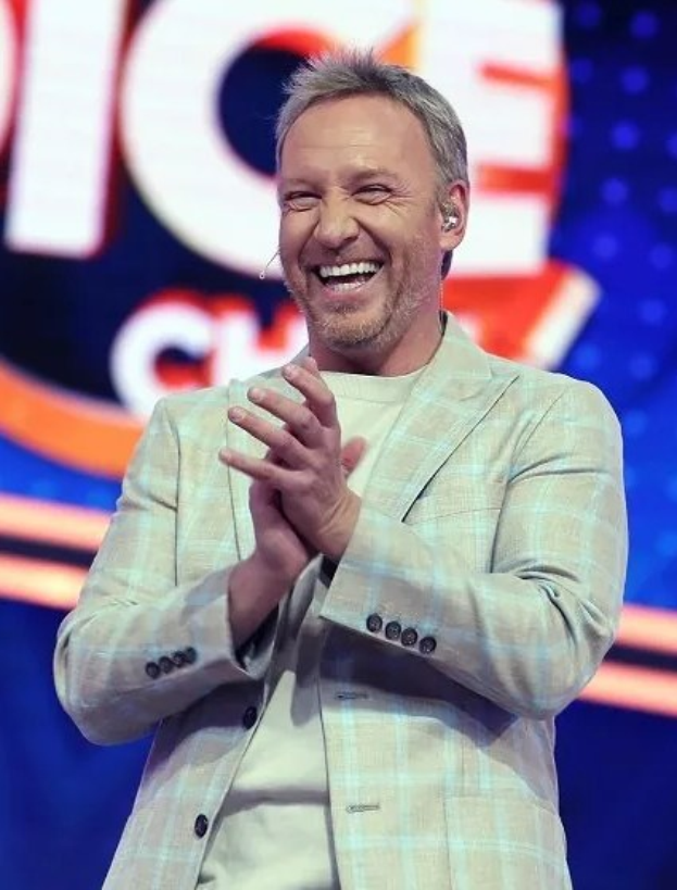 Ein Mann in einem Anzug, der auf einer Bühne klatscht, mit dem "The Voice"-Logo im Hintergrund.