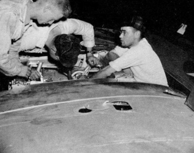 Drei Männer in Baseballkappen arbeiten an einem schwarz-weißen Auto in einer Garage, umgeben von Werkzeugen und Reparaturmaterialien.