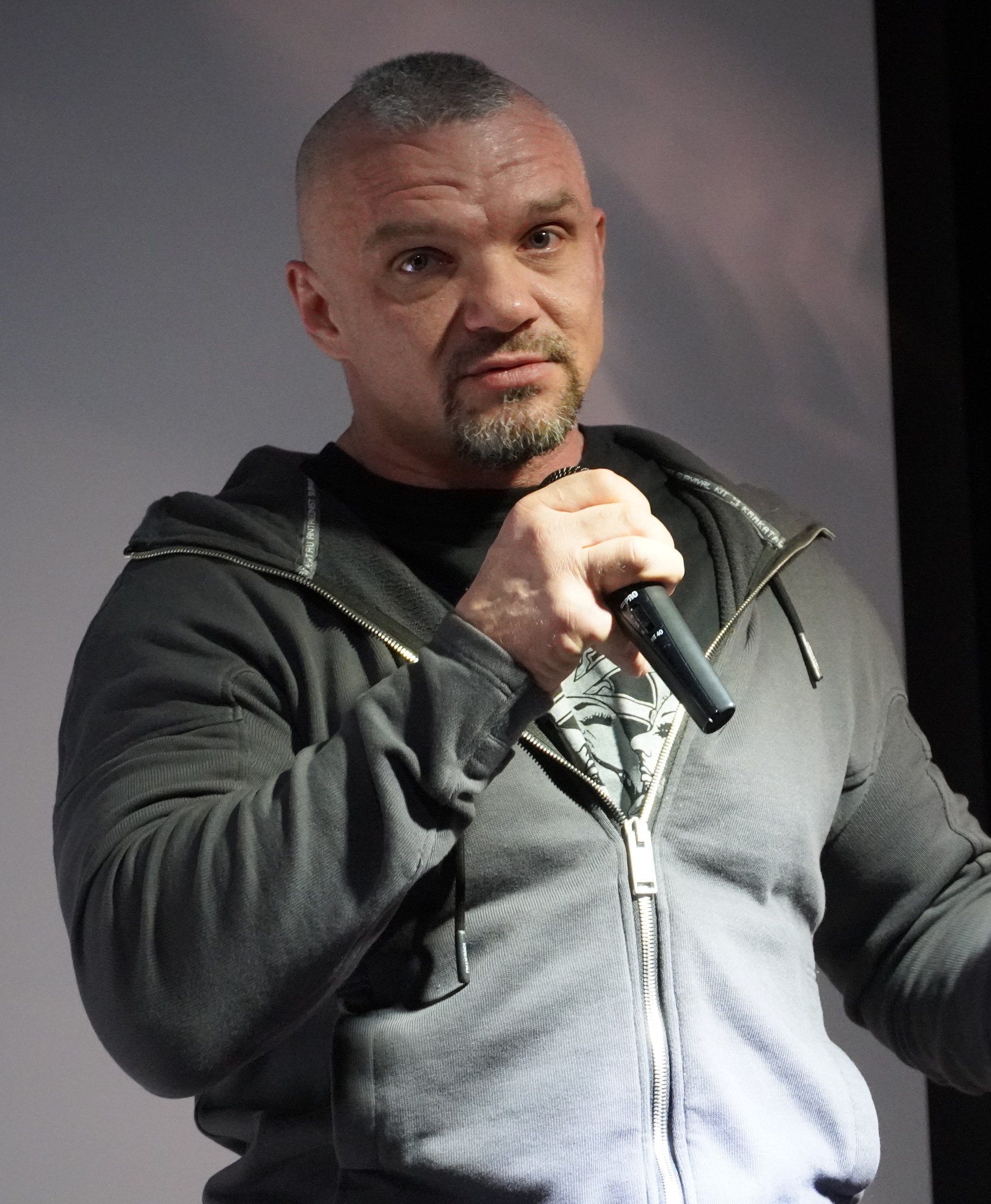 Ein Mann in einer schwarzen Kapuzenjacke und einem schwarzen Anzug mit einem weißen Hemd, glattem Haar und einem selbstbewussten Ausdruck h├Ąlt ein Mikrofon, waehrend er vor einer Wand steht.