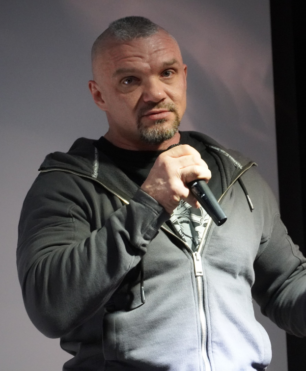 Ein Mann in einer schwarzen Kapuzenjacke und einem schwarzen Anzug mit einem weißen Hemd, glattem Haar und einem selbstbewussten Ausdruck h├Ąlt ein Mikrofon, waehrend er vor einer Wand steht.