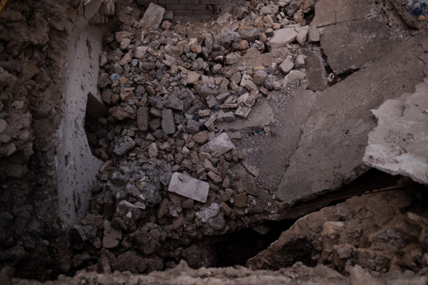 Zusammengebrochenes Gebäude mit einem großen Loch im Boden, umgeben von Trömmern und einer sichtbaren Leitung auf der linken Seite, die Erdbebenschäden anzeigt.