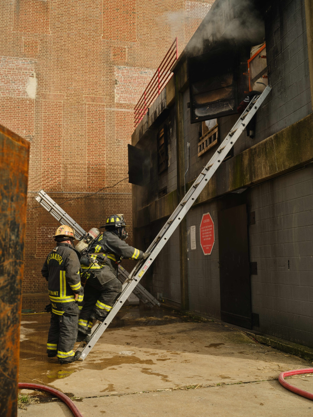 Feuerwehrleute in Helmen, die eine Leiter zu einem raucherfüllten Gebäude hinaufsteigen, mit Metallobjekten links und Rohren auf dem Boden.