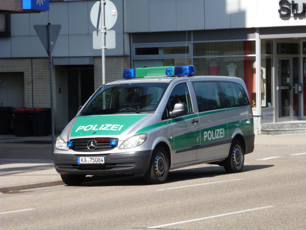 Ein Polizeiwagen parkt am Straßenrand vor einem Gebäude mit Glastüren, Schaufensterpuppen, Schildern und Mülltonnen im Hintergrund.