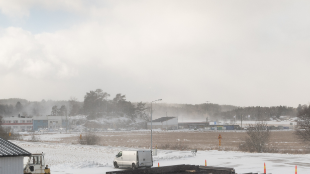Ein großer Lastwagen parkt tief im Schnee neben einem Gebäude, umgeben von Bäumen, Strommasten, Straßenlaternen und verstreuten Gegenständen unter einem bewölkten Himmel.