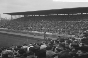 Schwarzes und weißes Foto eines vollen Fußballstadions mit Zuschauern, die ein Spiel verfolgen, mit Bannern, Pfählen, einem Schuppen, Bäumen, einem Turm und einem bewölkten Himmel.