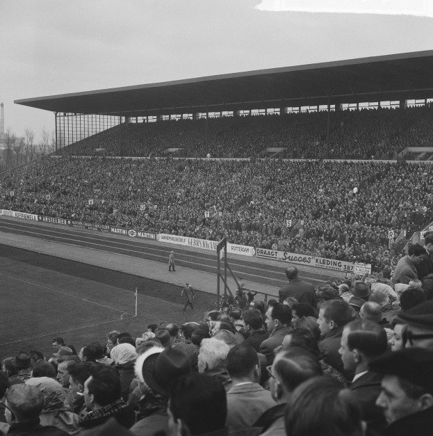 Schwarzes und weißes Foto eines vollen Fußballstadions mit Zuschauern, die ein Spiel verfolgen, mit Bannern, Pfählen, einem Schuppen, Bäumen, einem Turm und einem bewölkten Himmel.