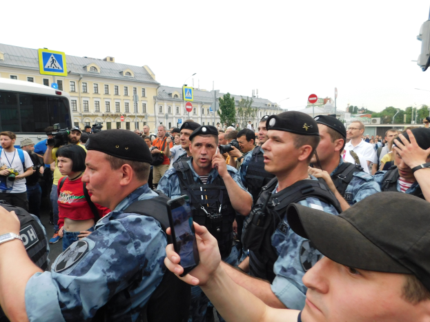 Eine Gruppe von Polizisten steht vor einer Menge von Menschen, die Kameras und Handys halten, mit einem Bus auf der linken Seite, Gebäuden und Bäumen im Hintergrund und einem klaren blauen Himmel.
