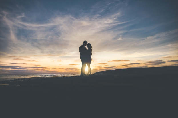 Silhouette eines Paares, das sich auf dem Strand bei Sonnenuntergang umarmt und küsst, mit einem bunten Himmel und tiefblauem Wasser im Hintergrund.