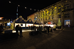 Ein lebendiger Weihnachtsmarkt in Berlin, Deutschland, mit Menschen um beleuchtete und geschmückte Stände, Gebäuden mit Fenstern im Hintergrund, unter einem dunklen Himmel.