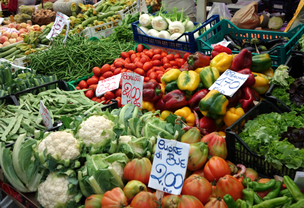 Ein belebter Bauernmarkt mit verschiedenen Gemüsen wie Tomaten, Paprika, Blumenkohl und grünen Bohnen in Körben, Preisetiketten sichtbar und einige Menschen anwesend.