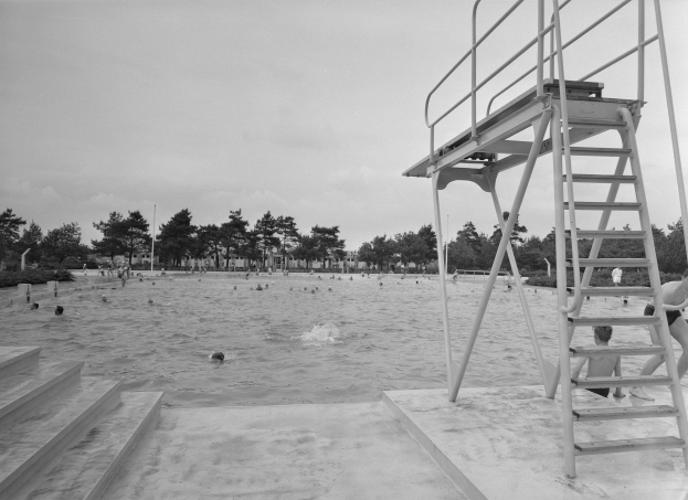 Schwarz-weißes Foto von Menschen beim Schwimmen an einem Strand mit einem Rettungsturm auf der rechten Seite mit Treppen, Bäumen und Pfählen im Hintergrund unter einem klaren Himmel.