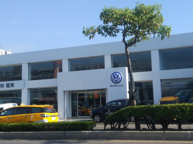Ein VW-Autohaus mit Glaswänden und -fenstern, das Autos zeigt, die vor dem Gebäude geparkt sind, umgeben von Pflanzen und einem Baum unter einem sichtbaren Himmel.