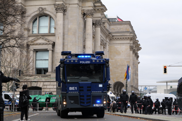 Gruppe von Polizisten vor einem großen Gebäude mit architektonischen Details, Fahrzeuge auf der Straße, eine Person mit einer Kamera auf der linken Seite, Bäume und Verkehrszeichen im Hintergrund unter einem klaren blauen Himmel.