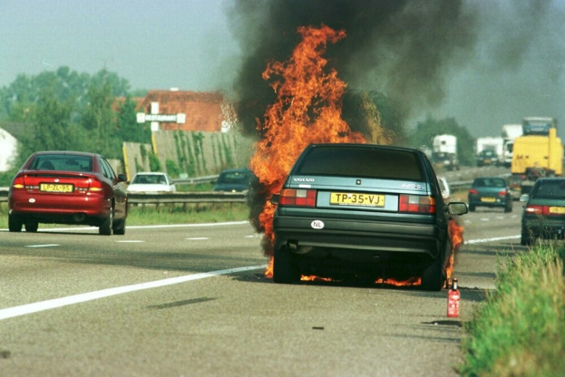 Ein Auto, das in Flammen steht, auf der Straße umgeben von anderen Fahrzeugen, mit Bäumen, Gebäuden und einem klaren blauen Himmel im Hintergrund und Gras und einem Feuerlöscher auf der rechten Seite.