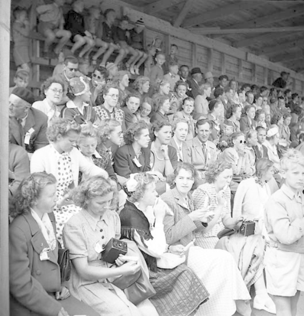 Schwarzes und weißes Foto einer Menge, die in einem Stadion sitzt, mit einer hölzernen Wand und einem Dach im Hintergrund, wobei einige Kameras halten.