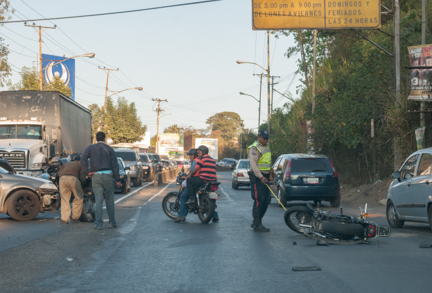Gruppe von Menschen um ein verunglücktes Motorrad auf dem Straßenrand versammelt mit mehreren Fahrzeugen, einschließlich eines Lastwagens, im Hintergrund und Bäumen, Pfählen, Lampen, Schildern und Himmel.