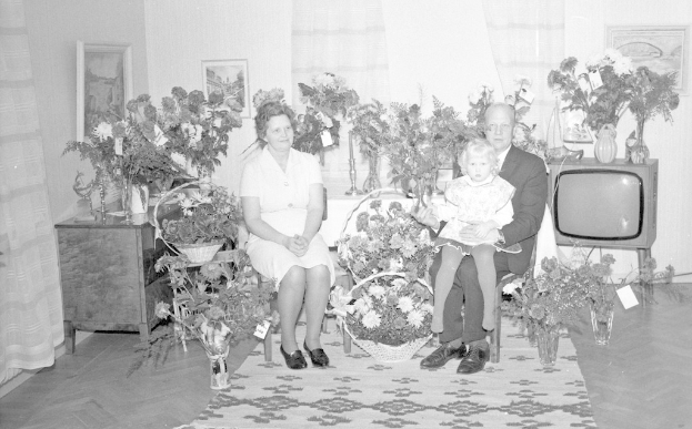 Schwarzes und weißes Foto einer Familie aus drei Personen - einem Mann, einer Frau und einem Kind - auf einem Sofa im Wohnzimmer mit Blumenvasen, einem Teppich, gerahmten Wanddekorationen und Vorhängen.