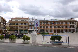 Ein belebter Stadtplatz mit Menschen, die sitzen und stehen, Topfpflanzen, Metallabsperrungen, ein Schild an einem Pfahl, Straßenlaternen mit Fahnen, Gebäude mit Fenstern und einem bewölkten Himmel.