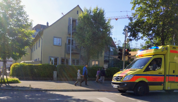 Ambulanz am Straßenrand mit Menschen auf dem Gehweg, Ampeln, Pfosten, Bäume, Pflanzen, Gras, Gebäude mit Fenstern und einen Kran im Hintergrund unter einem klaren blauen Himmel.