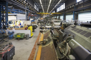 Großes Lagerhaus-Innere mit diverser Maschinen, einem Landfahrzeug auf der rechten Seite, metallischen Objekten auf der linken Seite und Deckenbeleuchtung.