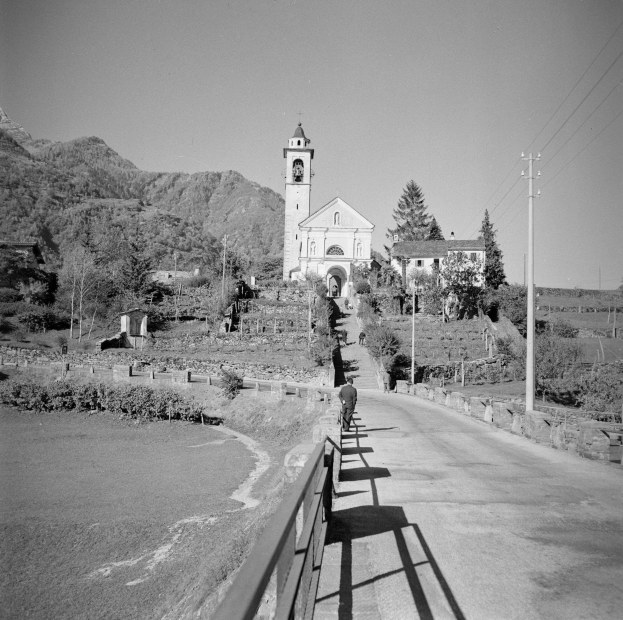 Schwarzes und weißes Foto einer Person, die ein Fahrrad auf einer Straße mit Geländern fährt, umgeben von Bäumen, Gebäuden, Masten, Drähten, H√∞geln und einem klaren Himmel.