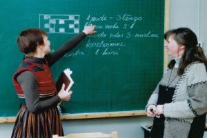 Zwei Frauen in einem Klassenzimmer, eine hält ein Buch und die andere schreibt an einer Tafel, mit Stühlen darunter.