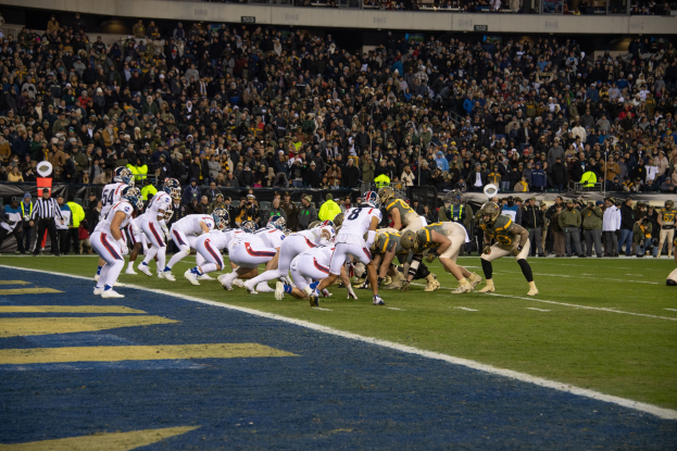 Ein Fußballspiel im Gange mit Spielern auf dem Boden in Sportbekleidung und Helmen, von einem Stadionplatz aus betrachtet.