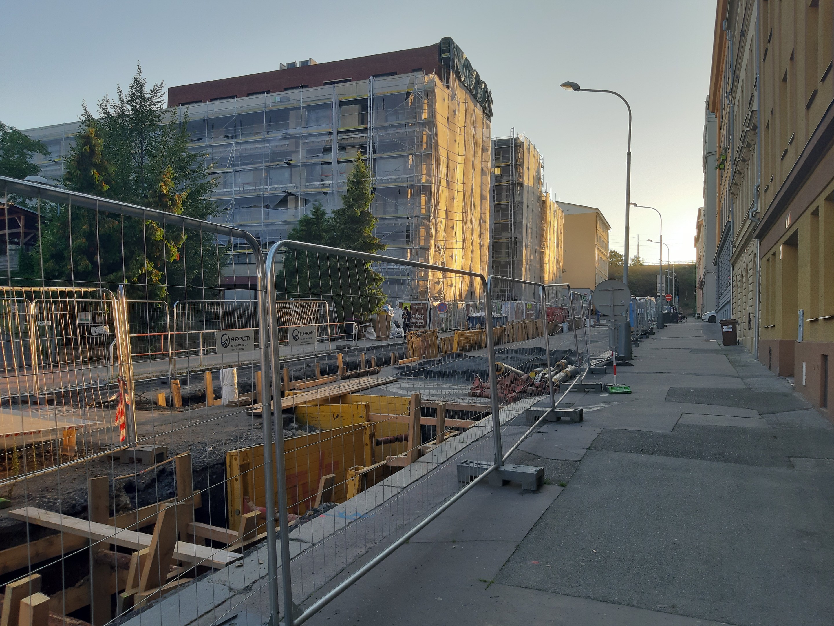 Eine Stadtstraße mit einer Baustelle in der Mitte, umgeben von Gebäuden, Straßenlaternen, Straßenlaternen, Fahrzeugen, Absperrpoller, Bäumen und einem klaren blauen Himmel.