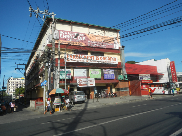 Eine belebte Stadtstraße mit einem großen Gebäude, Strommasten mit Kabeln, fahrenden Fahrzeugen, Fußgängern, einem Schirm, der Schatten spendet, Bäumen und einem klaren blauen Himmel im Hintergrund.
