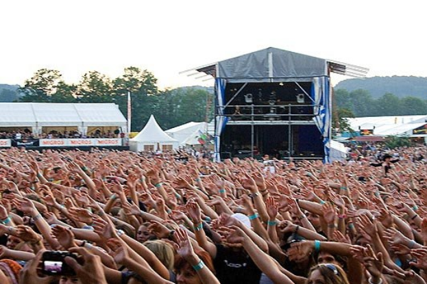 Menschenmenge auf einem Musikfestival mit erhobenen Händen und Handys, Bühne im Hintergrund unter Zelten und Bäumen mit blauem Himmel.