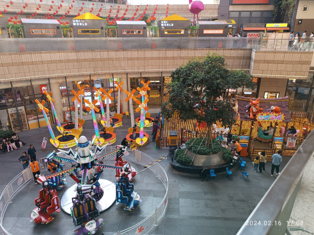 Ein Vergnügungspark innerhalb eines Einkaufszentrums mit Fahrgeschäften, Bäumen, Menschen, Stühlen und Buden, mit Gebäuden oben und einem Wasserzeichen unten rechts.