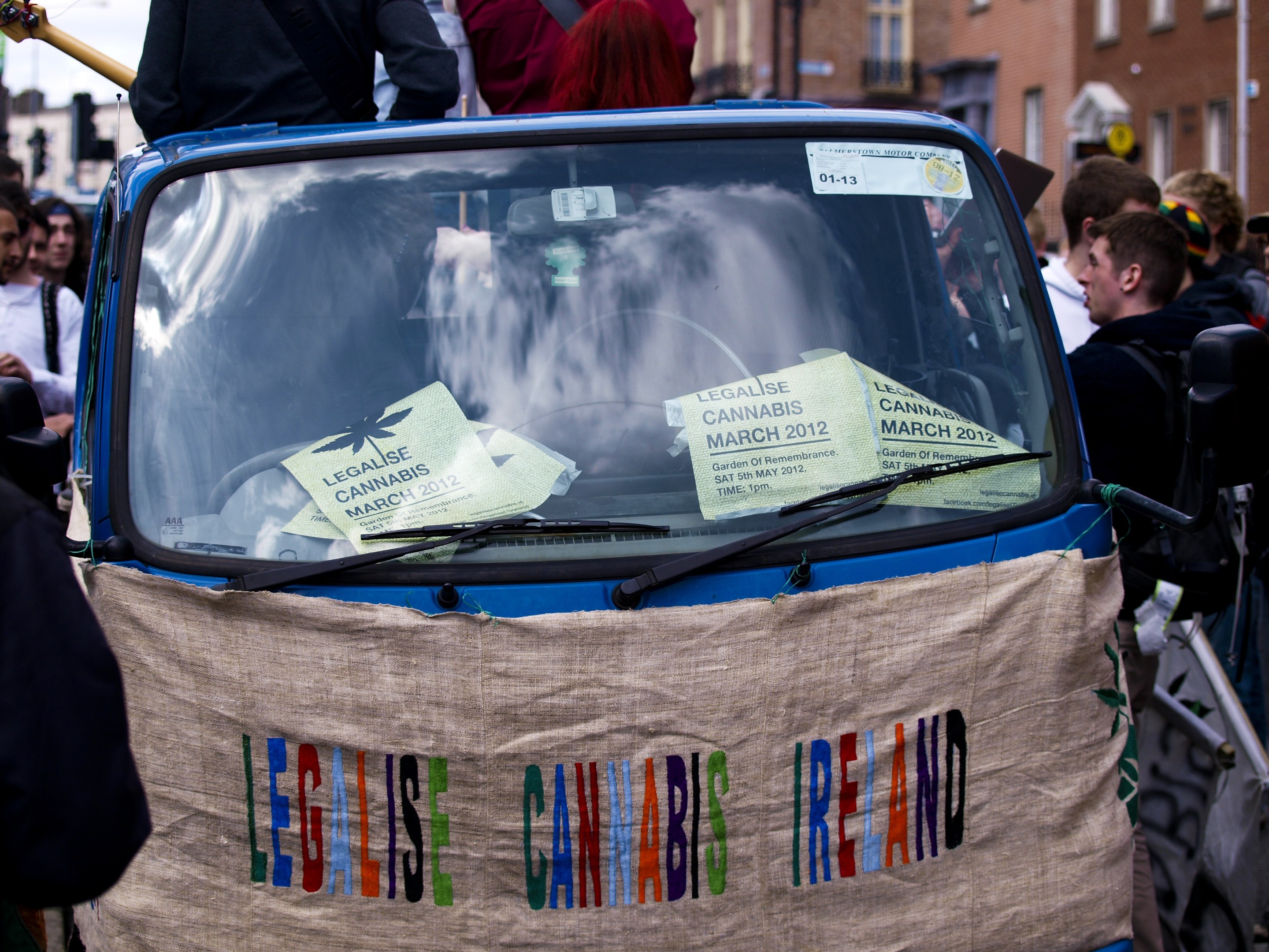 Gruppe von Menschen um ein Auto mit einem "Legalise Cannabis Ireland"-Schild herumstehend, umgeben von Gebäuden und einem klaren blauen Himmel, mit Papieren im Auto sichtbar.