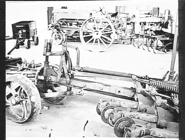 Schwarzes und weißes Foto eines Traktors in einer Fabrik, mit einem Pflug im Vordergrund und einer Wand im Hintergrund, eingerahmt von einem Rahmen.