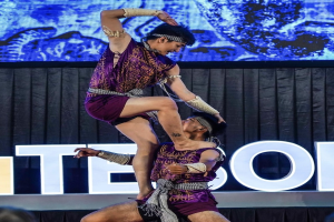 Zwei Performer in purpurfarbenen Kleidern führen einen akrobatischen Hebemanöver auf der Bühne vor einem Publikum aus, mit schwarzem Vorhang und Bildschirm im Hintergrund und sitzenden Zuschauern darunter.