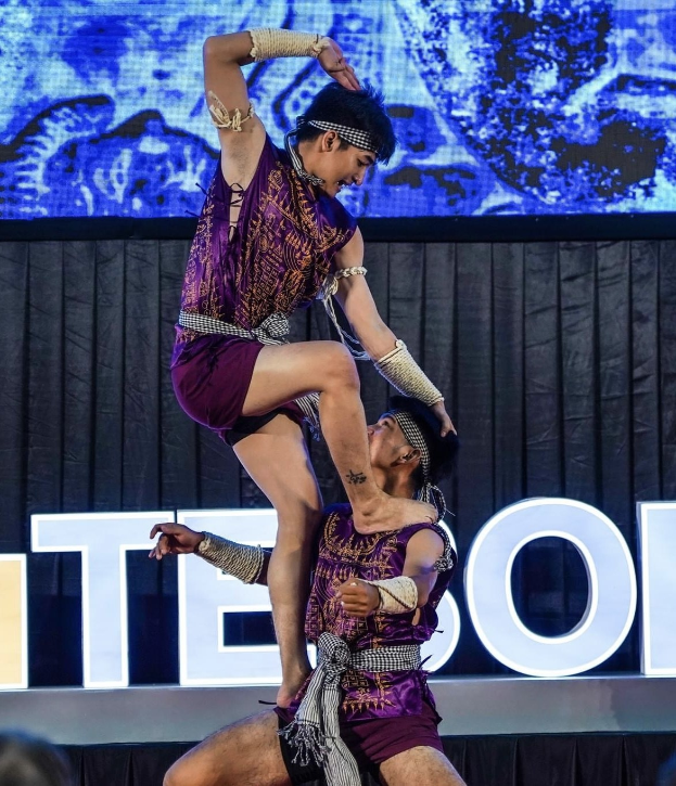 Zwei Performer in purpurfarbenen Kleidern führen einen akrobatischen Hebemanöver auf der Bühne vor einem Publikum aus, mit schwarzem Vorhang und Bildschirm im Hintergrund und sitzenden Zuschauern darunter.