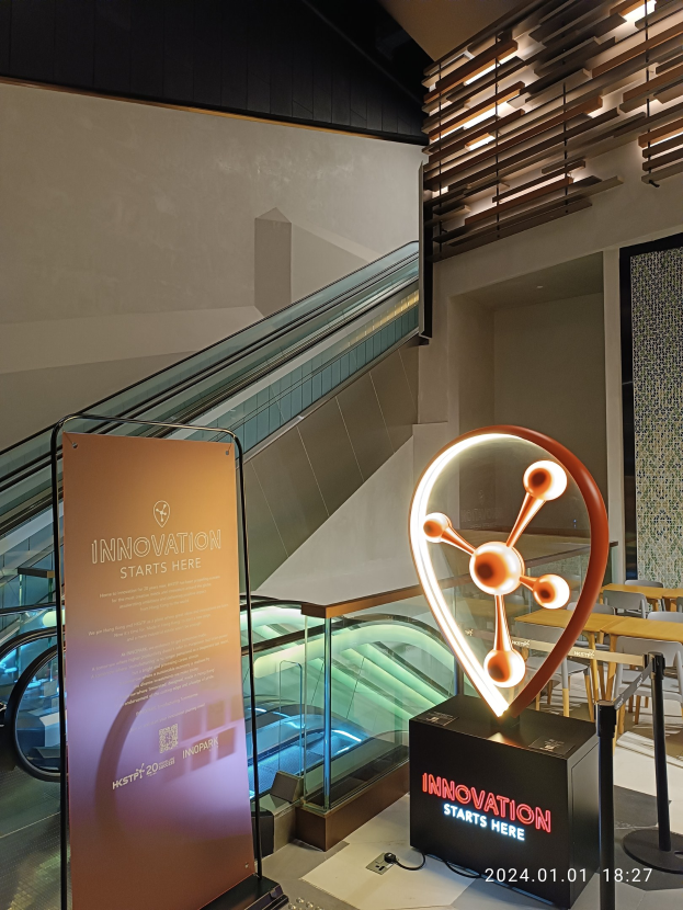 Großes Schild mit der Aufschrift "Innovation beginnt hier" in einem Gebäude mit einer Rolltreppe im Hintergrund, Tische und Stühle auf der rechten Seite und ein Schild, das auf die HSTP20 Expo hinweist.
