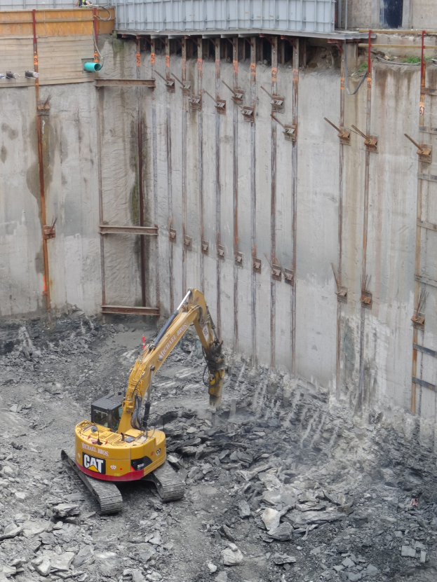 Ein Bagger ist auf einer Baustelle im Einsatz, umgeben von Steinen und Eisenstangen, mit einem teilweise errichteten Gebäude links im Bild sichtbar.