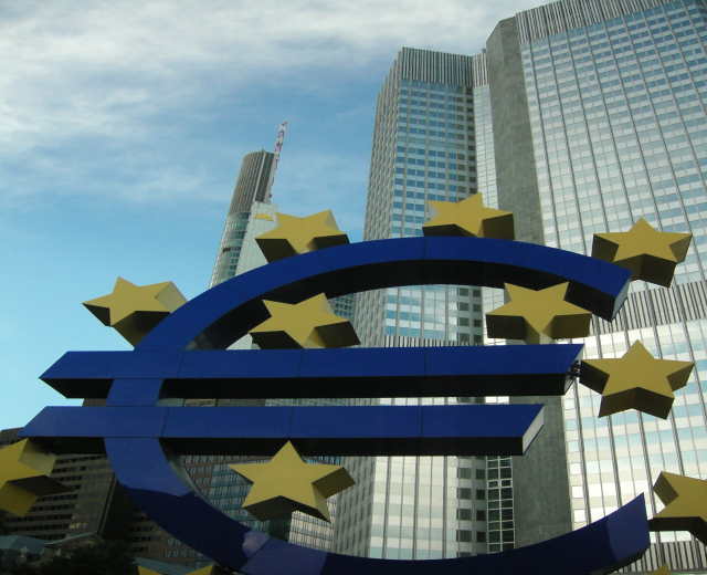 Eurozeichen vor dem Hauptsitz der Europäischen Zentralbank in Frankfurt, Deutschland, mit Bäumen und bewölktem Himmel im Hintergrund.