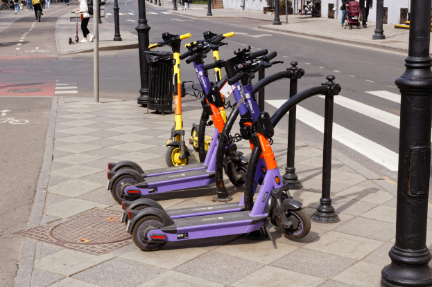 Eine Reihe von Elektro-Scootern, die entlang einer Straße mit Fußgängern, Radfahrern und einem Müllcontainer im Vordergrund und Gebäuden, Bäumen und Pflanzen im Hintergrund geparkt sind.