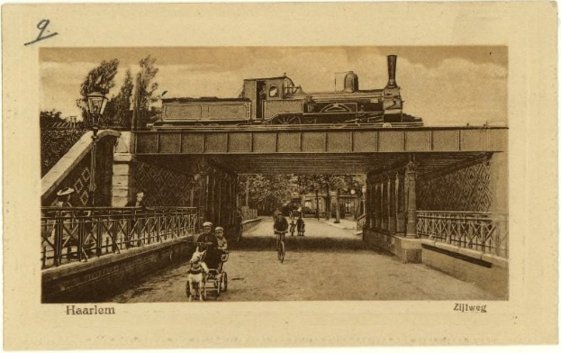 Altes Schwarz-Weiß-Foto einer Brücke, über die ein Zug fährt, mit Menschen auf Fahrrädern und einem Menschen in einem Kinderwagen, Bäume im Hintergrund und Text unten.