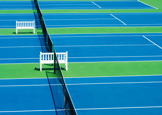 Ein blau-grüner Tennisplatz mit zwei weißen Bänken, einem Netz in der Mitte und umgeben von saftigem Grün.