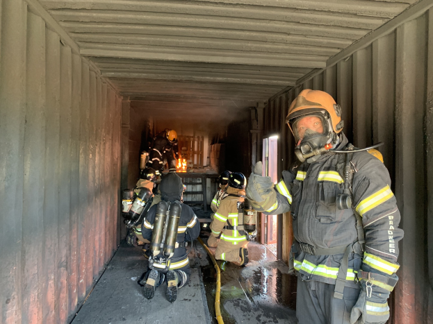 Feuerwehrleute in Schutzausru├čtung, einschlie├člich Helmen, Handschuhen und Sauerstoffflaschen, arbeiten innerhalb eines Seecontainers mit einem Feuerl├Âscher im Hintergrund.
