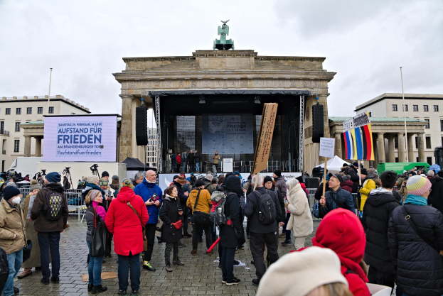 Eine Menschenmenge steht vor einem Gebäude mit einer Bühne, Lautsprechern und einem Bildschirm und hält Fahnen und Schilder, was auf eine Protestaktion in Berlin hindeutet.