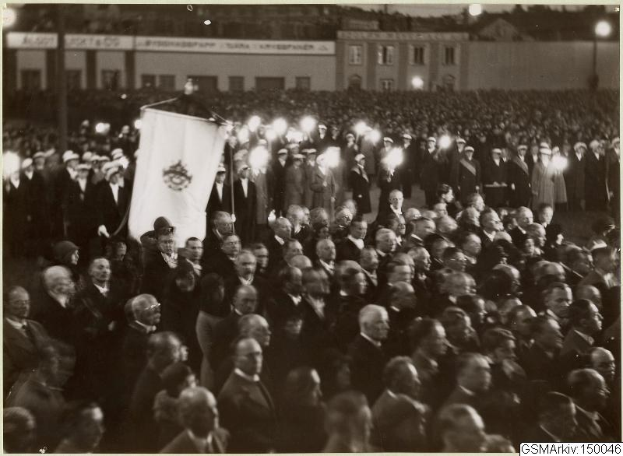 Schwarzes und weißes Foto einer großen Menge vor einem Gebäude, mit einer Person, die eine Fahne hält, und Text am unteren Rand: "Gustav Klimts Rede an der Berliner Mauer."
