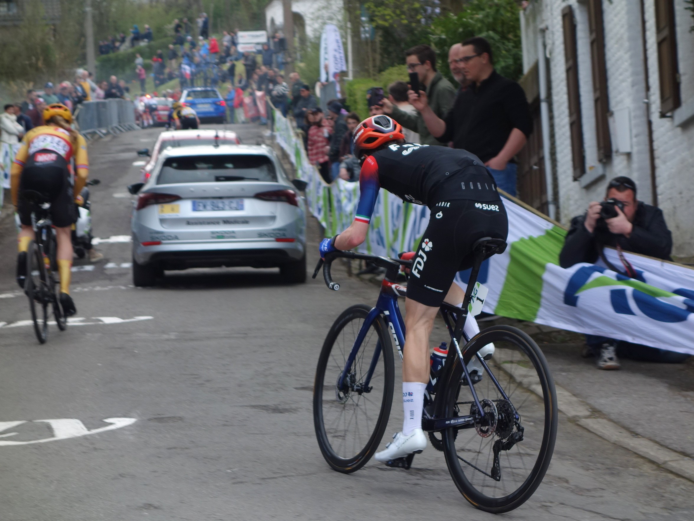 Eine Gruppe von Menschen, die auf Fahrrädern die Straße entlangfahren, mit Autos neben ihnen, Menschen mit Kameras und einer Fahne auf der rechten Seite und einem Zaun, Bäumen, Gebäuden und Himmel im Hintergrund.