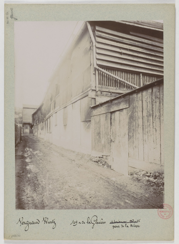 Ein altes Schwarz-Weiß-Foto einer Schotterstraße, die von ein paar Gebäuden gesäumt ist, mit Text am unteren Rand des Bildes.