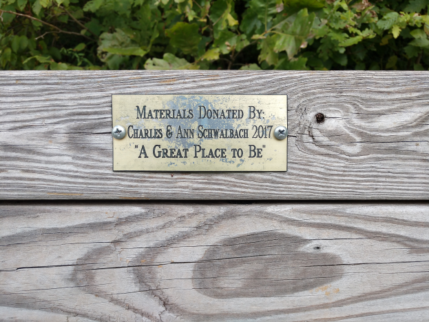 Eine hölzerne Bank mit einer Plakette, auf der "Materialien gespendet von Charles & Ann Schwabach 2017: Ein toller Ort" steht, umgeben von üppigen grünen Pflanzen.